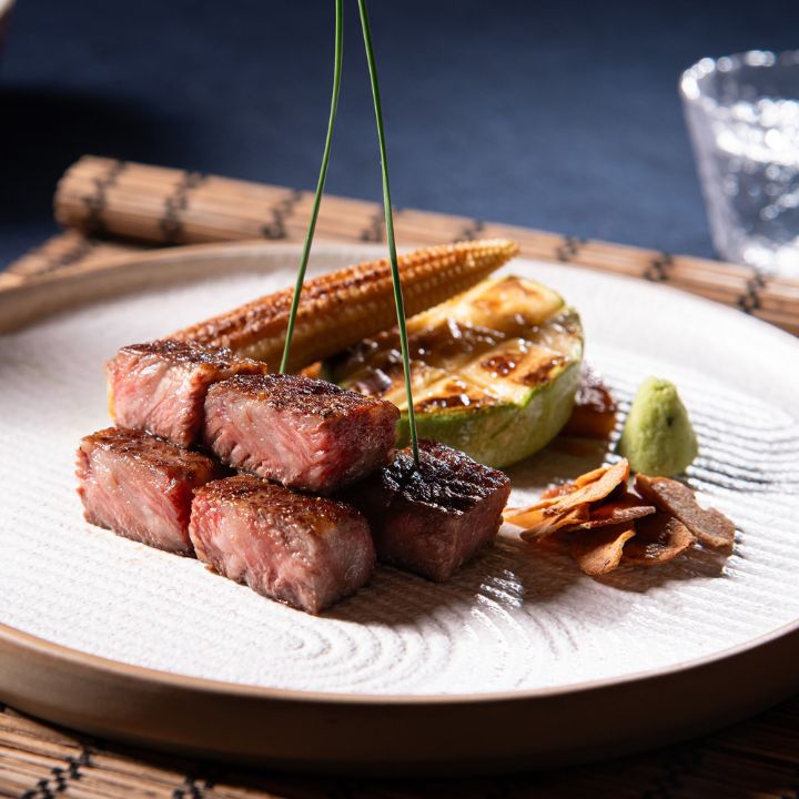 Wagyu beef cubes, grilled zucchini and baby corn served in ceramic dish on bamboo placemat against blue background.